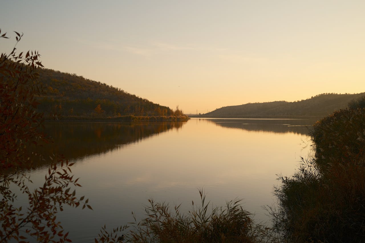 A tranquil sunset on a calm lake surrounded by hills and lush foliage, creating a serene landscape.